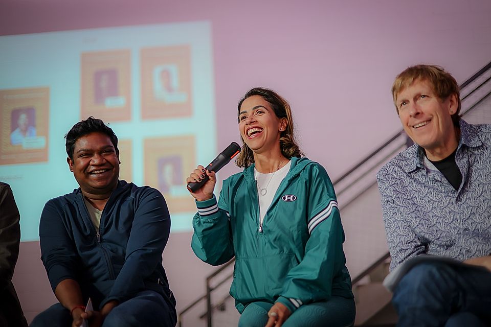 Three speakers at a panel discussion, one holding a microphone, smiling, with a presentation screen behind them.