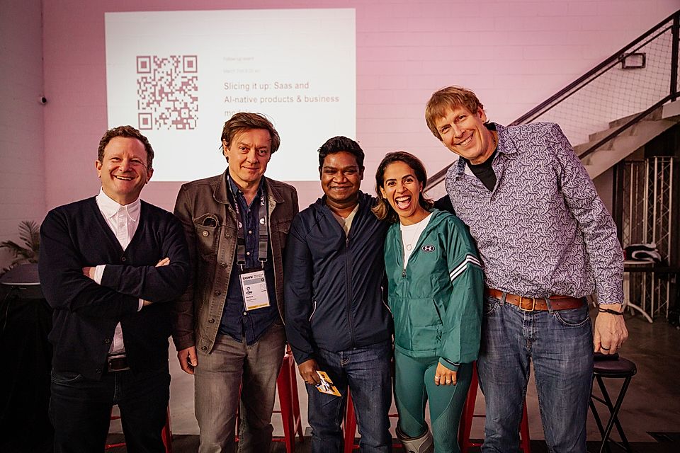 Five professionals posing together at an event, with a presentation screen in the background and casual attire.