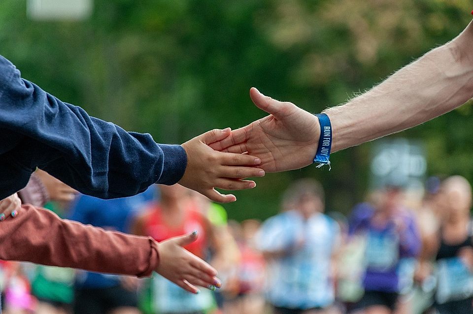 Two hands reaching out to connect during a running event, with blurred runners in the background.