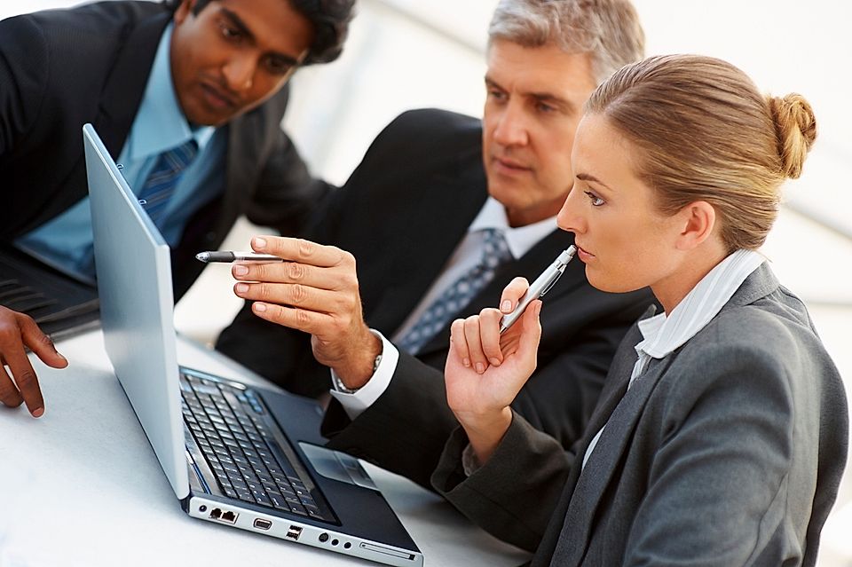 Three business professionals discussing on a laptop during a meeting in a modern office setting.