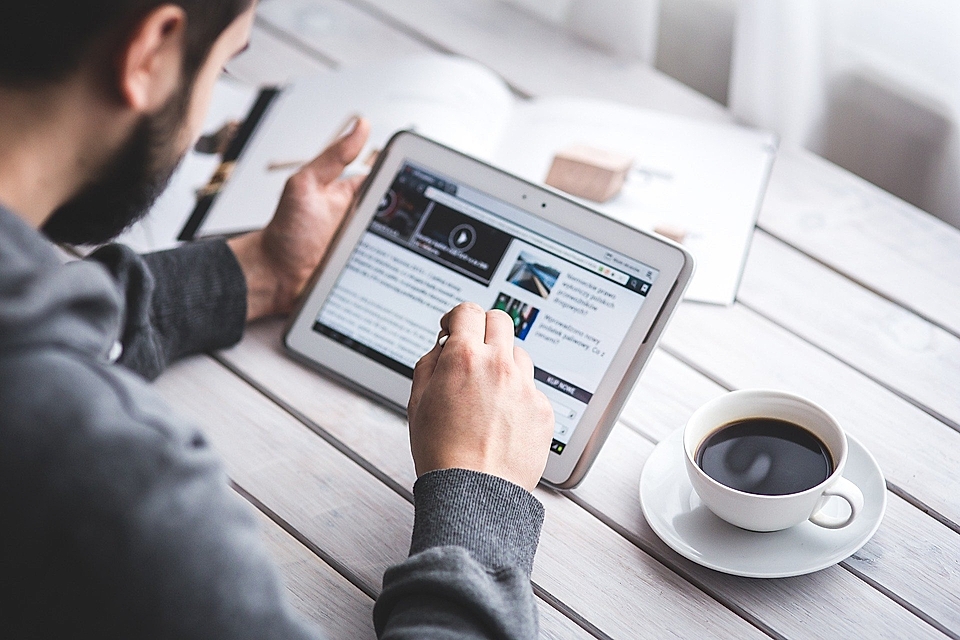 A person using a tablet to browse content, with a cup of coffee on a wooden table. Magazine pages visible in the background.