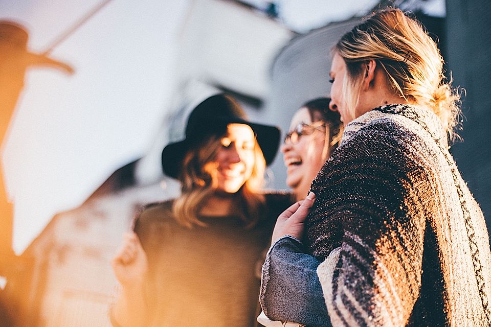 Three women laughing together outdoors, wearing casual clothing, with soft sunlight creating a warm atmosphere.