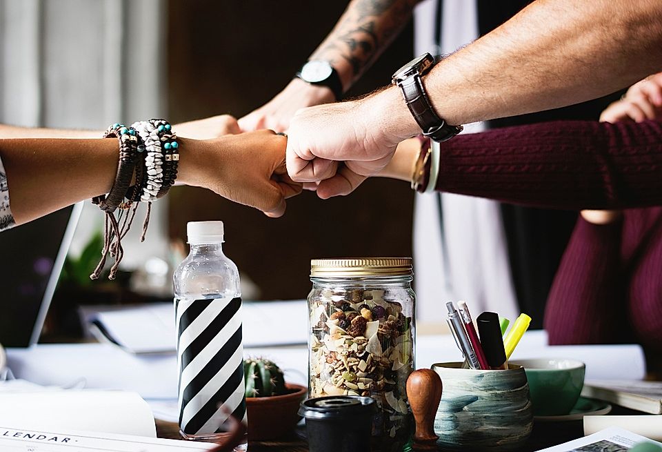 Hands of diverse individuals fist-bumping over a table with a water bottle, a jar of snacks, and office supplies.