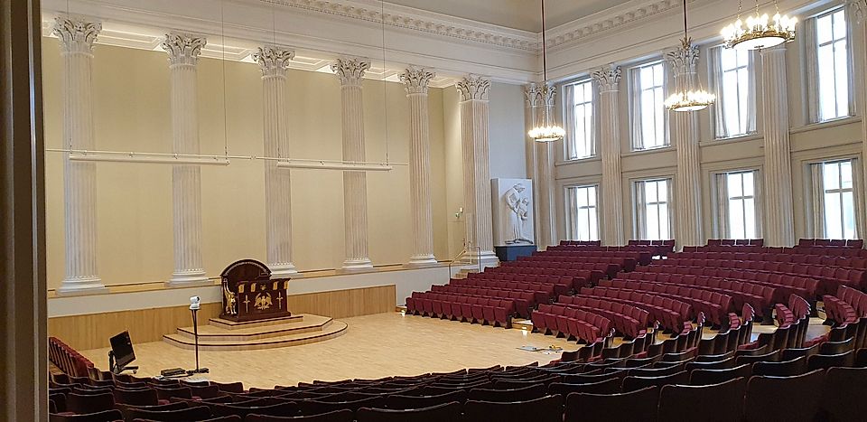 Spacious auditorium with red seating, tall columns, and a central lectern, featuring large windows and bright lighting.
