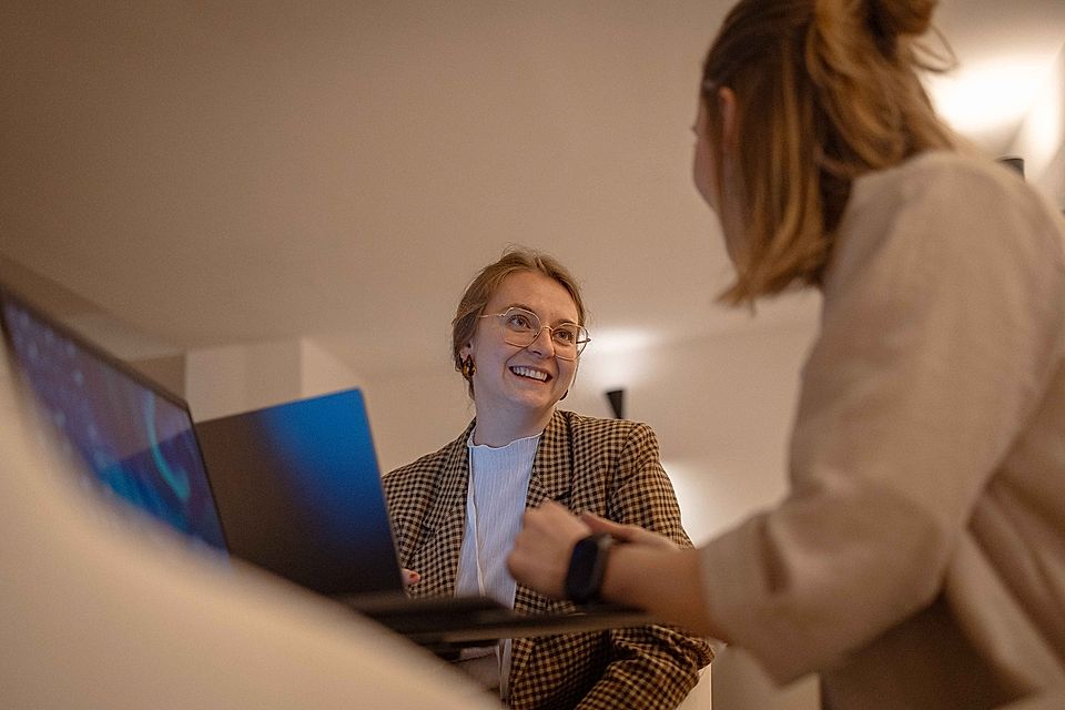 Two women in a modern office setting discussing work, with a laptop on the table, one smiling and wearing glasses.