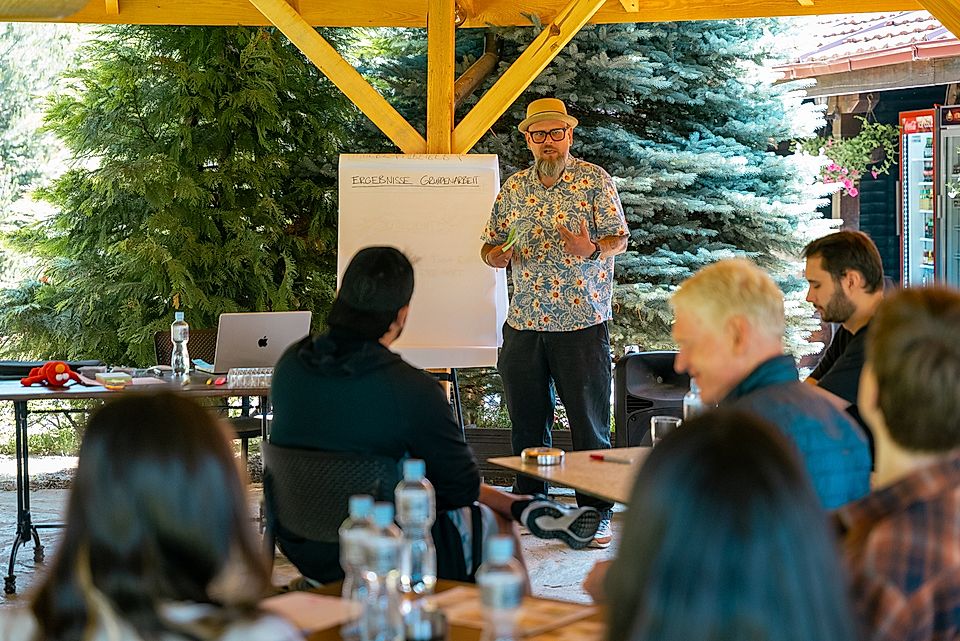 A speaker presents in an outdoor workshop setting, with participants seated at tables, engaged in discussion.