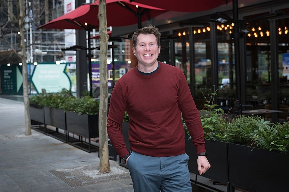 Smiling man in a red sweater standing outdoors beside a café with planters and string lights.