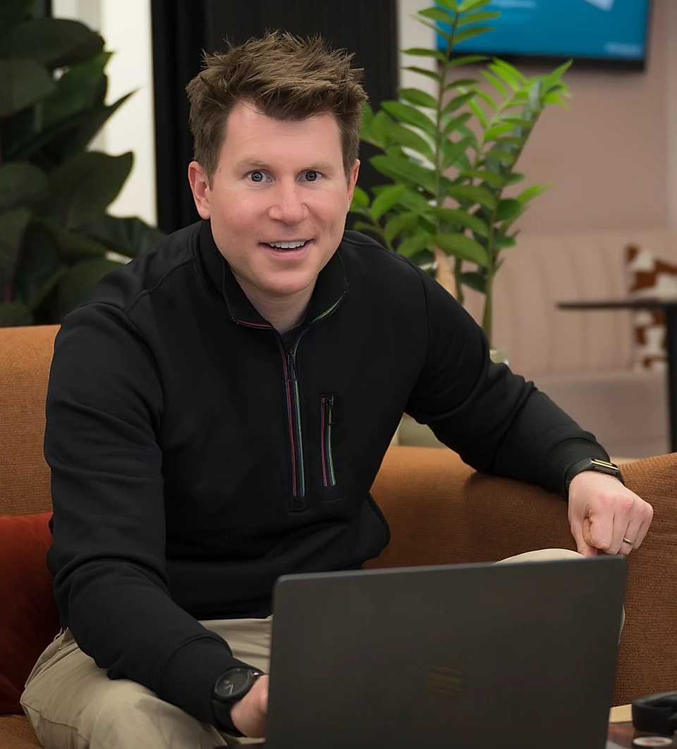 Man with short hair, wearing a black pullover, smiles while sitting on a couch and using a laptop in an office setting.