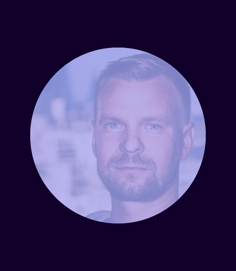 A headshot of a man with light brown hair and a beard, set against a blurred city background.