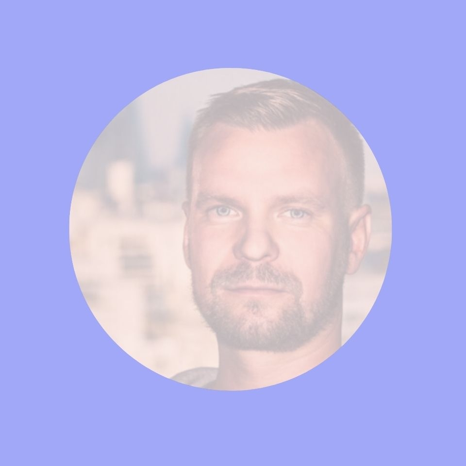 A man with a beard and short hair, wearing a light shirt, smiles against a blurred city background.