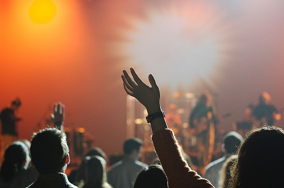 Audience with hands raised at a concert, colorful lights in the background, and musicians performing on stage.