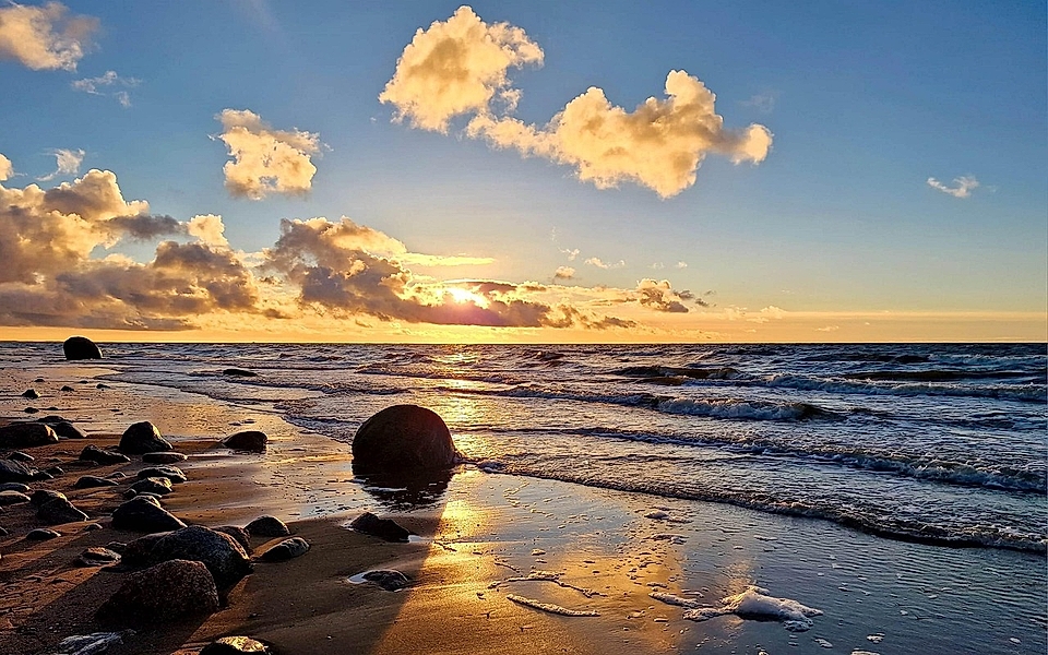 Sunset over the ocean, with waves lapping against a sandy beach and large rocks, under a blue sky with clouds.
