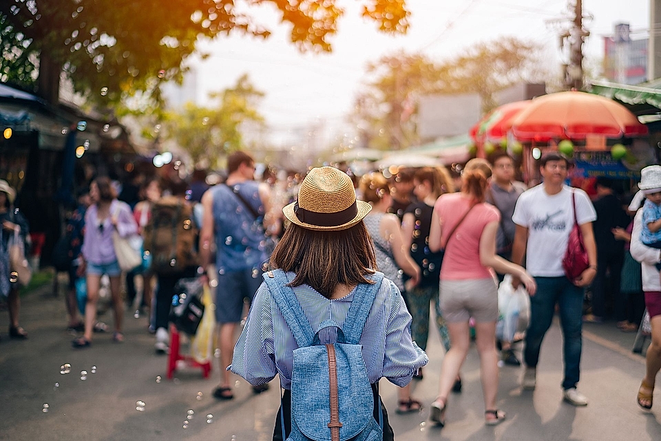 A woman in a hat stands in a busy market street filled with people, colorful stalls, and bubbles in the air.