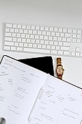 Flat lay of a white keyboard, planner with handwritten notes, and a gold wristwatch against a white background.