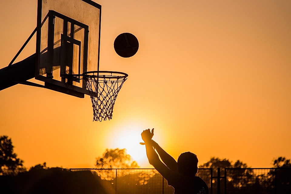 Silhouette of a person shooting a basketball towards a hoop at sunset, with vibrant orange and yellow sky.