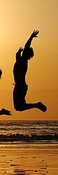 Silhouette of a person jumping joyfully on the beach at sunset, with ocean waves and a warm, orange sky.