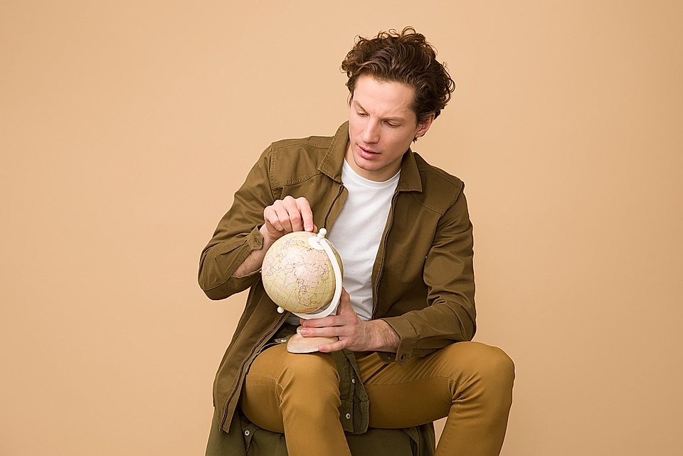 A young man in a brown jacket examines a globe, seated against a beige background.