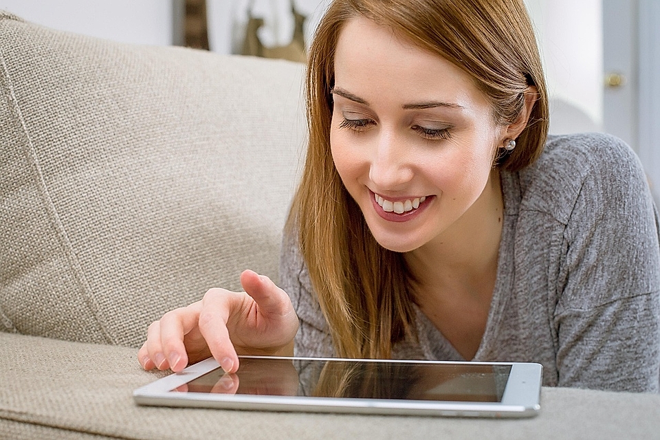 A woman with long hair smiles while using a tablet on a sofa, focusing on the screen, in a bright living room.
