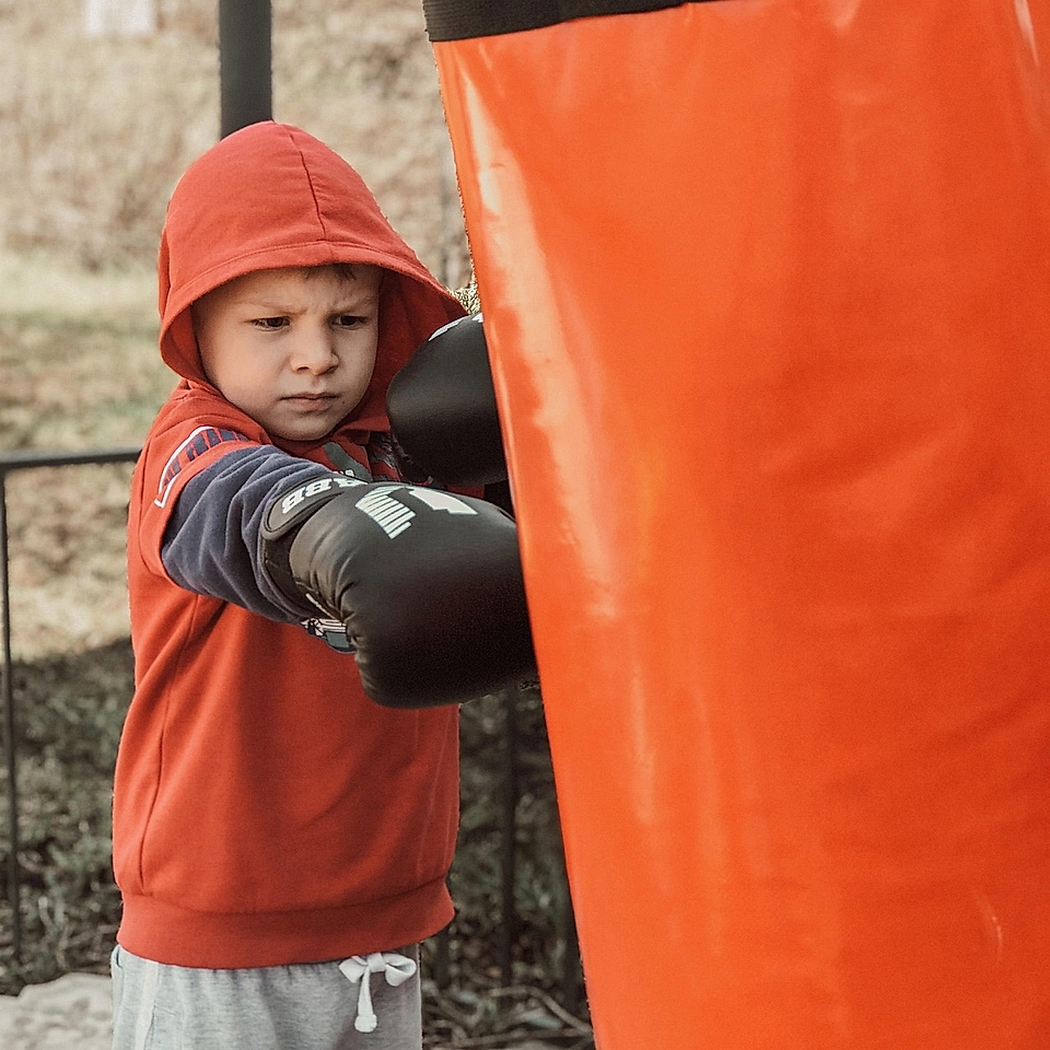 A young boy in a red hoodie boxing against an orange punching bag, wearing black boxing gloves, focused on his stance.