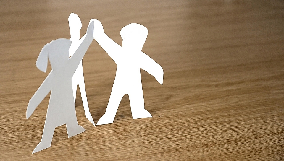 Three paper cutout figures, two standing and one raised hand, on a wooden surface.