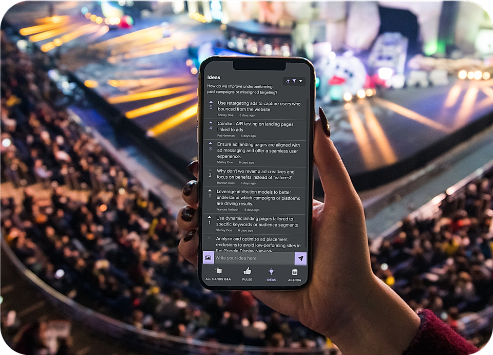 A person holds a smartphone displaying marketing ideas at a crowded event with stage lights in the background.