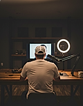 Person in a cap sitting at a desk, facing a monitor with a webcam ring light, in a dimly lit room.