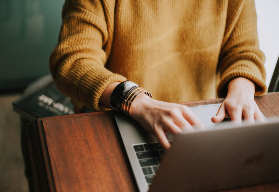 A person in a mustard sweater typing on a laptop while wearing multiple bracelets, seated at a wooden desk.