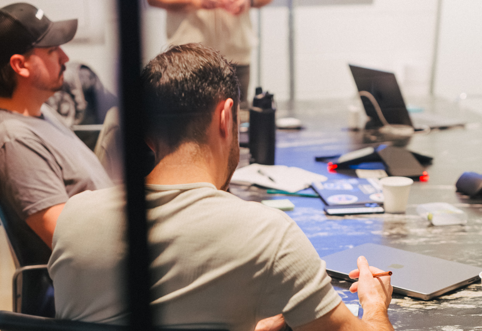 Two men in a meeting, engaged in discussion at a table covered with laptops, notebooks, and a coffee cup.