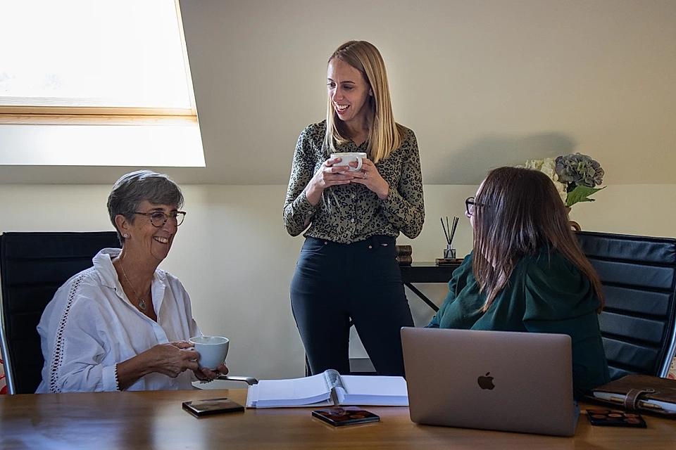 Three women talking in a meeting room with laptops, notebooks, and coffee cups