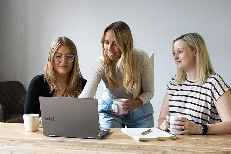 Three women collaborating at a table with a laptop, notebook, and coffee mugs in a meeting room.