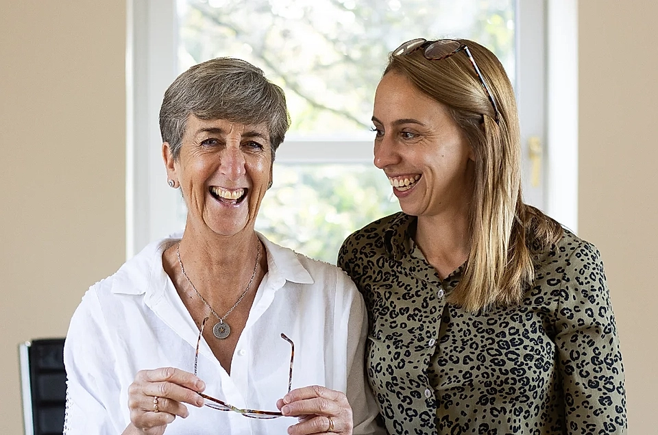 Two smiling women standing together indoors, one holding eyeglasses, in front of a bright window.