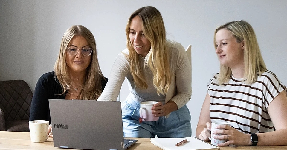 Three women gathered around a laptop at a table, discussing work and holding mugs in a bright office.