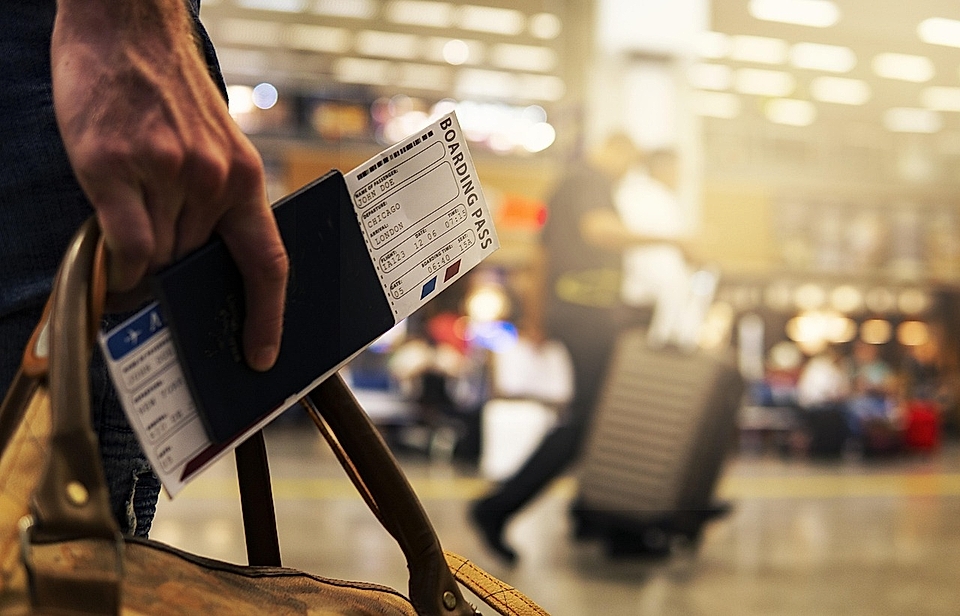 A traveler holding a passport and boarding pass at an airport, with luggage and blurred passengers in the background.