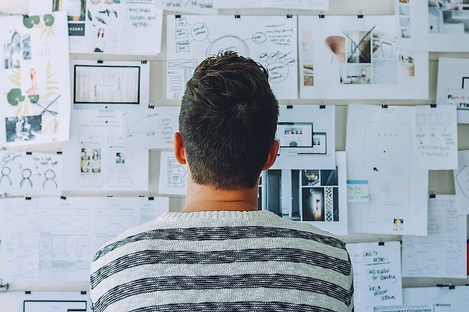 A person viewed from behind, looking at a wall filled with various papers, notes, and design mockups.