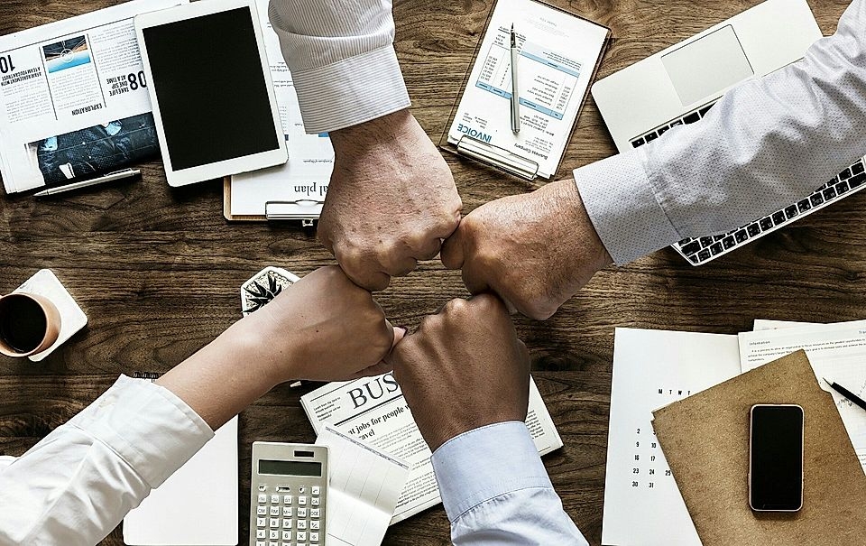 Four hands fist-bumping over a wooden desk with papers, a calculator, and electronic devices, symbolizing collaboration.