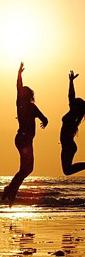 Silhouettes of two women jumping joyfully at the beach during sunset, with waves gently lapping at the shore.