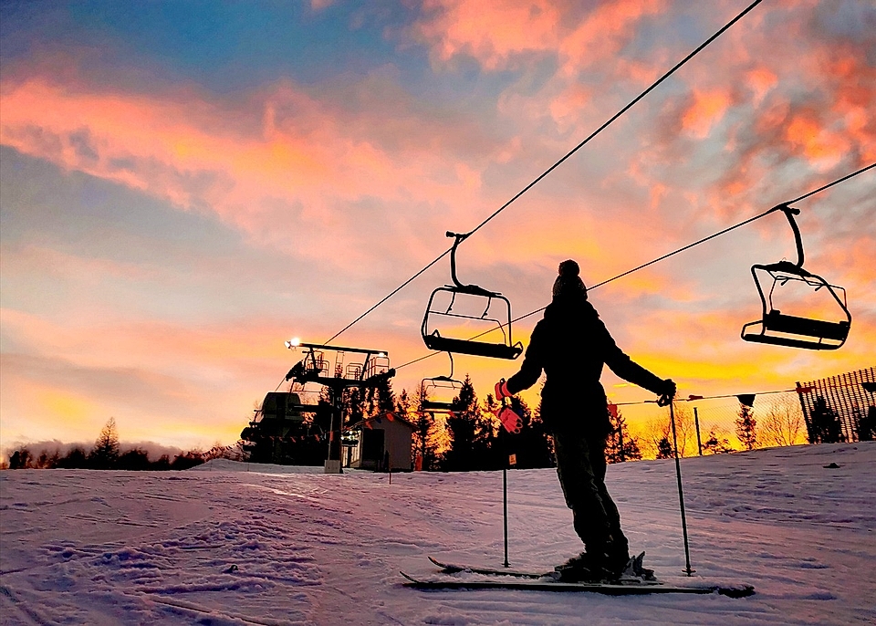 A skier stands on a snowy slope at sunset, with ski lifts in the background and colorful clouds above.