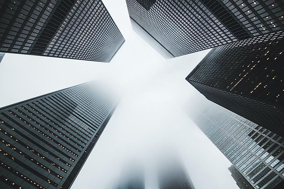 Low-angle view of modern skyscrapers shrouded in fog against a cloudy sky.
