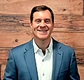 A smiling man in a blue blazer and white shirt stands in front of a wooden wall, looking directly at the camera.