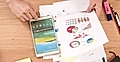 A tablet displaying data alongside printed graphs and charts on a wooden desk, with office supplies nearby.