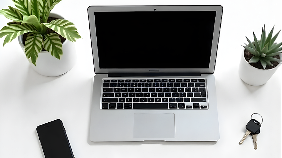 Laptop on a white surface with a smartphone, keys, and two potted plants in the background.