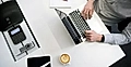 An overhead view of a person typing on a laptop, with a coffee cup, notepad, and office phone on the desk.