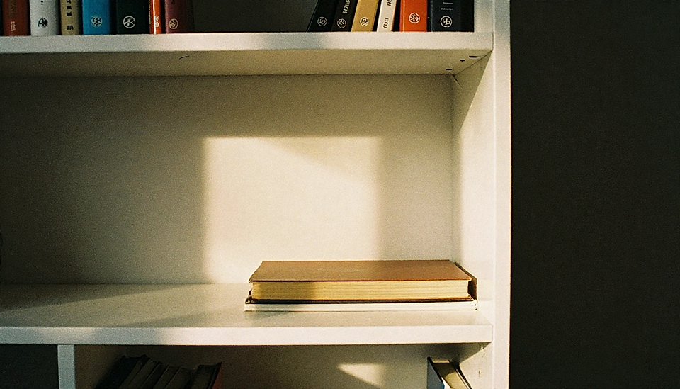 A white bookshelf holds a stack of books, with one open book illuminated by natural light casting a shadow.