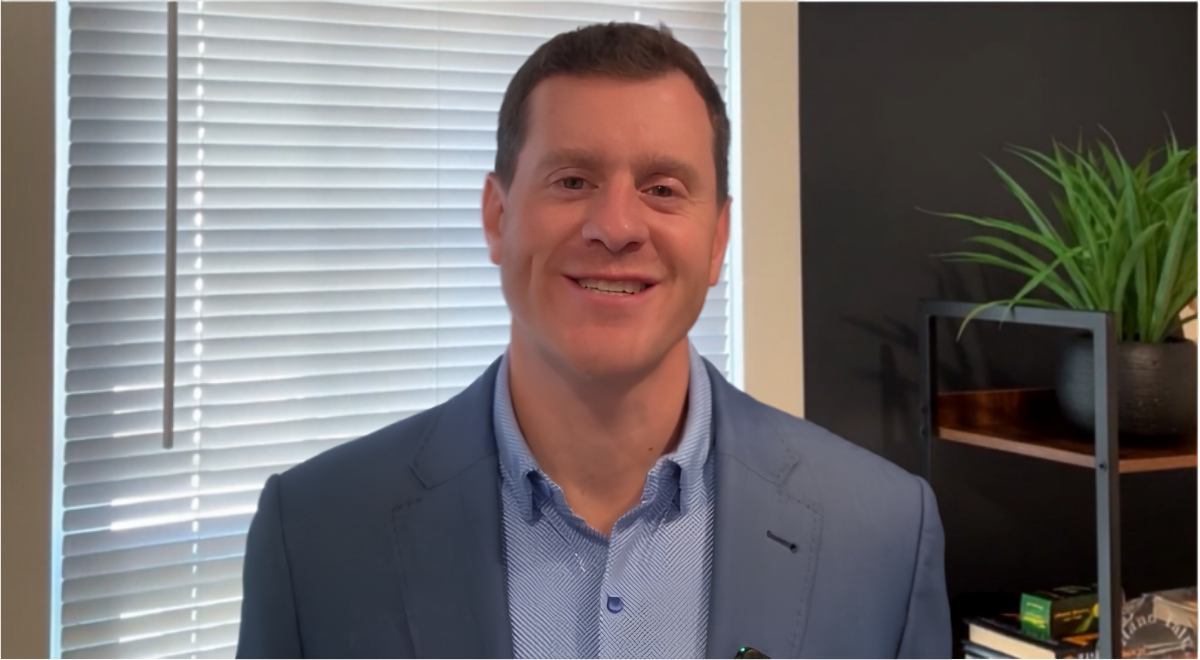 Smiling man in a blue blazer standing indoors beside window blinds and a plant
