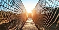 Suspension bridge with mesh sides illuminated by sunlight, leading toward a wooden platform in the distance.