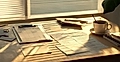 A sunlit desk with papers, a clipboard, a pen, and a coffee cup, featuring shadows from nearby plants.