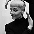 A person with short, light-colored hair poses creatively with arms raised, wearing a black turtleneck, in black and white.