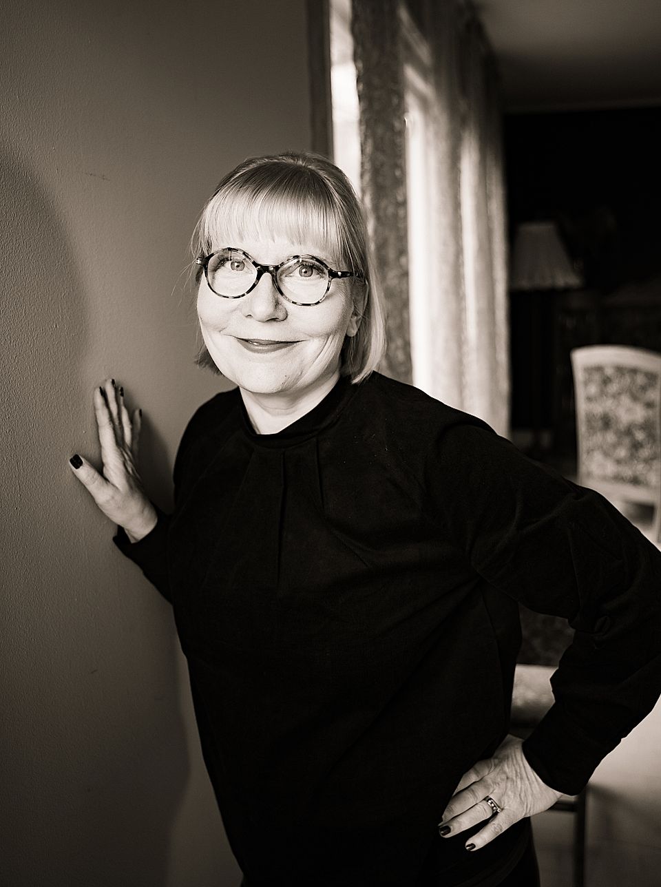 Smiling woman with round glasses and black top posing indoors by a wall in black-and-white portrait