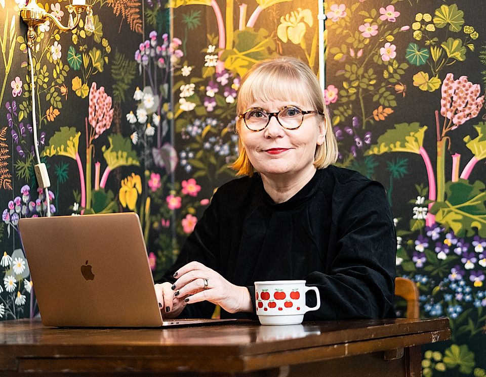 A woman with glasses sits at a wooden table, using a laptop. A colorful floral mural decorates the background.