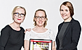 Three women in professional attire pose together, holding a laptop displaying a website, against a white background.